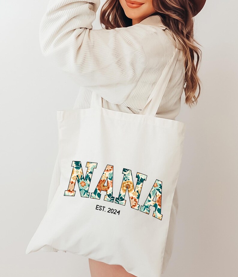 Op de afbeelding: Witte canvas draagtas met het woord "NANA" in grote, gedurfde letters. De letters zijn gevuld met een bloemenpatroon in tinten oranje, geel, groen en blauw. De tekst "EST. 2024" is onder het woord "NANA" gedrukt.