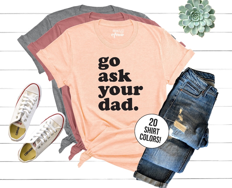 May include: A peach colored t-shirt with the text "go ask your dad." in black block letters. The shirt is folded and displayed with a pair of blue jeans and white sneakers.