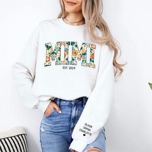 May include: White sweatshirt with the word "MIMI" in floral print letters and the text "EST. 2024" below. The sweatshirt has a black heart and the names "Oliver", "Sophia", and "Charlotte" printed on the left sleeve.