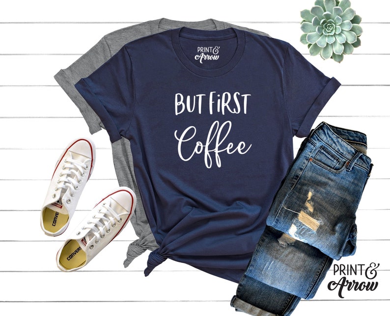 May include: A navy blue t-shirt with the text "But First Coffee" in white lettering. The shirt is laid out on a white wooden surface with a pair of white trainers and a pair of blue jeans.