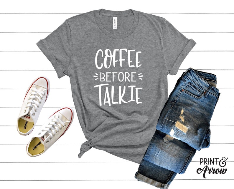 May include: A heather gray t-shirt with the text "Coffee Before Talkie" in white lettering. The shirt is laid out on a white wooden surface with a pair of white sneakers and a pair of blue jeans.