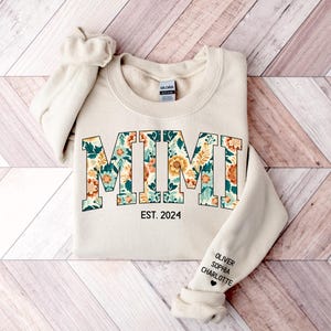 May include: A beige sweatshirt with the word "MIMI" in large, floral letters. The letters are outlined in black and filled with a floral pattern in shades of green, orange, and yellow. Below the word "MIMI" is the text "EST. 2024". The sleeve of the sweatshirt has the names "OLIVER, SOPHIA, CHARLOTTE" written in black.