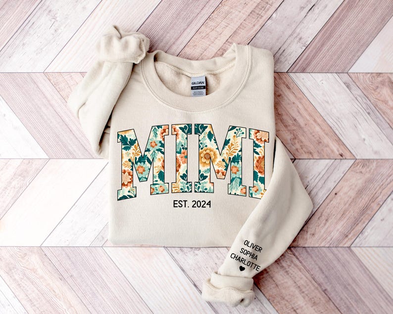 May include: A beige sweatshirt with the word "MIMI" in large, floral letters. The letters are outlined in black and filled with a floral pattern in shades of green, orange, and yellow. Below the word "MIMI" is the text "EST. 2024". The sleeve of the sweatshirt has the names "OLIVER, SOPHIA, CHARLOTTE" written in black.
