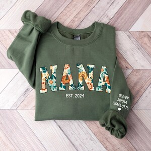 Op de afbeelding: Een groen sweatshirt met het woord "NANA" in bloemenprint letters. De tekst "EST. 2024" is gedrukt onder het woord "NANA". De namen "OLIVER", "SOPHIA" en "CHARLOTTE" zijn gedrukt op de rechtermouw van het sweatshirt.