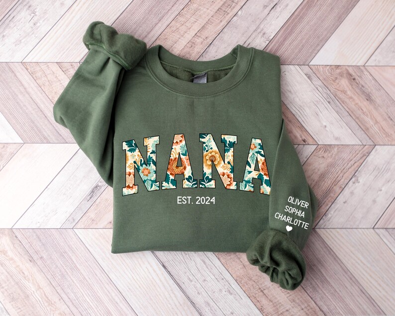 Op de afbeelding: Een groen sweatshirt met het woord "NANA" in bloemenprint letters. De tekst "EST. 2024" is gedrukt onder het woord "NANA". De namen "OLIVER", "SOPHIA" en "CHARLOTTE" zijn gedrukt op de rechtermouw van het sweatshirt.
