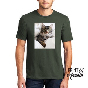 Puede incluir: Una camiseta de manga corta y cuello redondo en color verde oliva oscuro con un estampado gr&aacute;fico rectangular de un gato atigrado de pelo largo con ojos verdes descansando sobre ropa de cama blanca. El texto "PRINT & Arrow" aparece en negro.