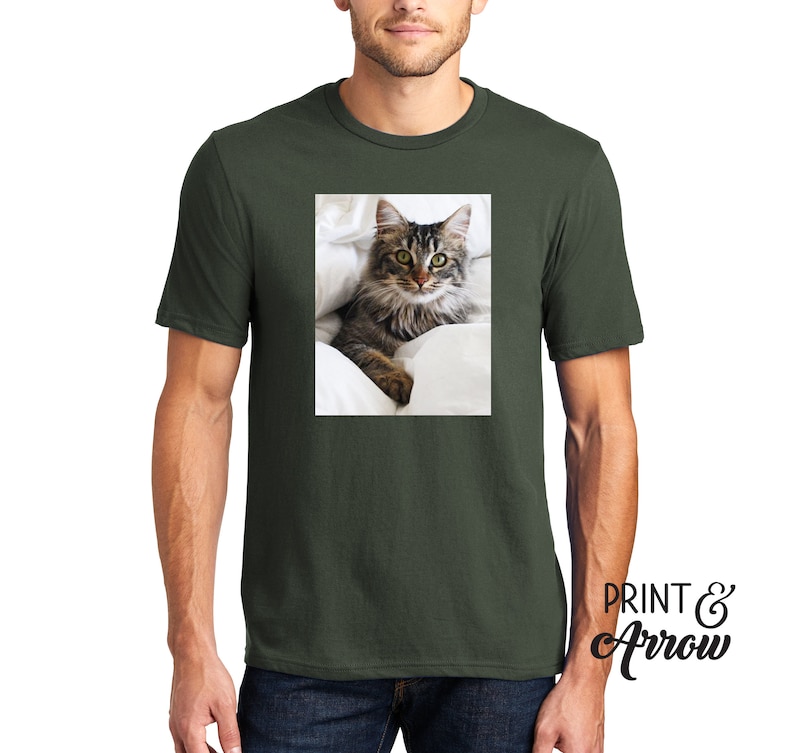 Puede incluir: Una camiseta de manga corta y cuello redondo en color verde oliva oscuro con un estampado gr&aacute;fico rectangular de un gato atigrado de pelo largo con ojos verdes descansando sobre ropa de cama blanca. El texto "PRINT & Arrow" aparece en negro.