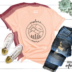 May include: A peach-colored t-shirt with a black graphic of a mountain range, sun, and birds inside a circle. The text "explore more" is written below the circle. The t-shirt is laid out on a white wooden surface with a pair of white sneakers, a pair of blue jeans, and a succulent plant.