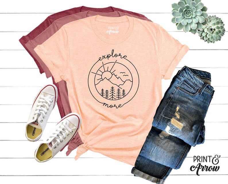 May include: A peach-colored t-shirt with a black graphic of a mountain range, sun, and birds inside a circle. The text "explore more" is written below the circle. The t-shirt is laid out on a white wooden surface with a pair of white sneakers, a pair of blue jeans, and a succulent plant.