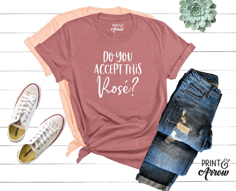 May include: A pink t-shirt with the text "Do you accept this ros&eacute;?" in white. The shirt is laid out on a white wooden surface with a pair of white sneakers, a pair of blue jeans, and a small potted succulent.