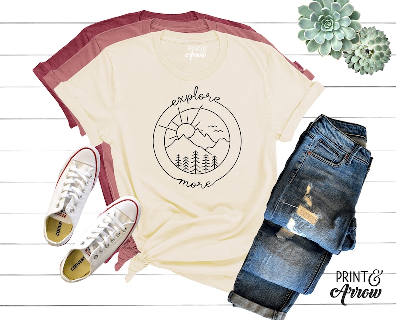 May include: A cream colored t-shirt with a black graphic of a mountain range, sun, and birds inside a circle with the text "explore more".