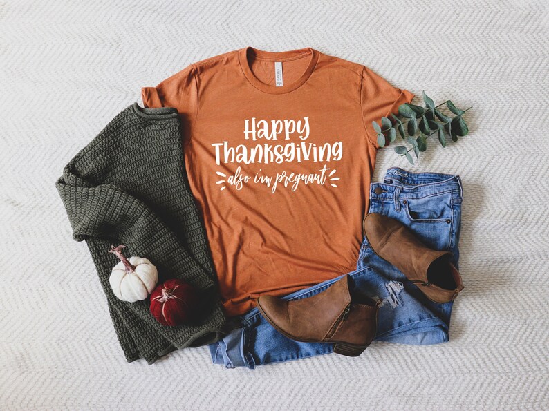 May include: A burnt orange t-shirt with the text "Happy Thanksgiving also I'm pregnant" in white lettering. The shirt is laid out on a white and grey chevron patterned surface with a green sweater, brown boots, and blue jeans.