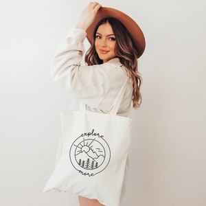 May include: A white canvas tote bag with a black line drawing of a mountain range, sun, and birds inside a circle. The text "explore more" is written below the circle.