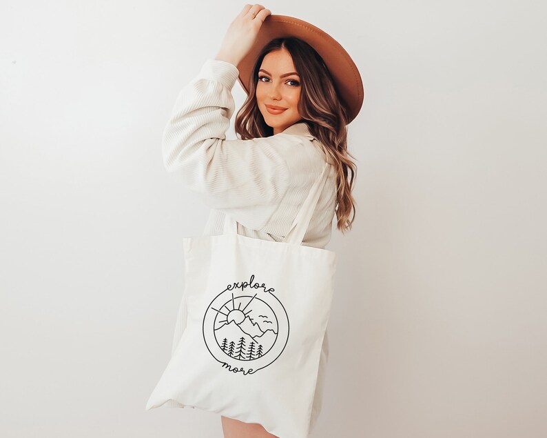 May include: A white canvas tote bag with a black line drawing of a mountain range, sun, and birds inside a circle. The text "explore more" is written below the circle.