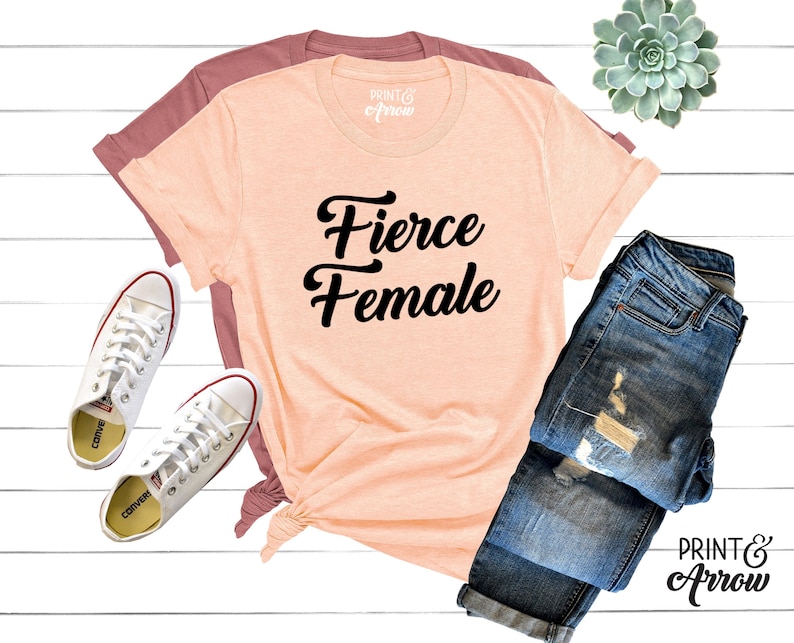May include: A peach-colored t-shirt with the text "Fierce Female" in black cursive lettering. The shirt is laid out on a white wooden surface with a pair of white sneakers, a pair of blue jeans, and a small potted succulent plant.