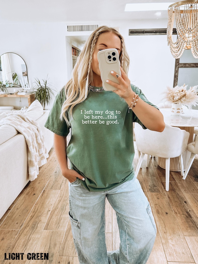 May include: Light green t-shirt with the text "I left my dog to be here...this better be good." The shirt is paired with light blue jeans. The image is taken indoors with a neutral background.