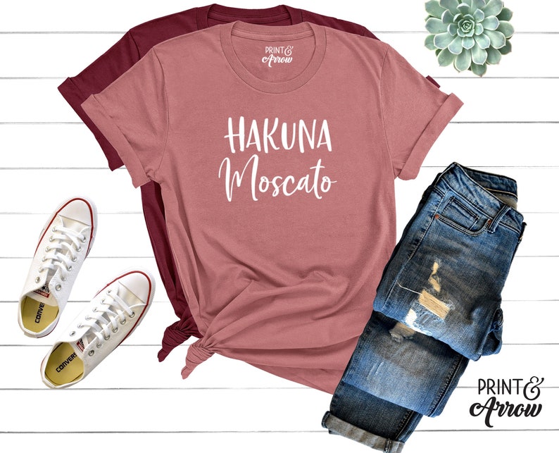 May include: A pink t-shirt with the text "Hakuna Moscato" in white lettering. The shirt is laid out on a white wooden surface with a pair of white sneakers, a pair of blue jeans, and a small green succulent plant.
