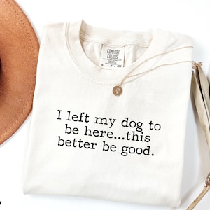 May include: Ivory-colored t-shirt with the text "I left my dog to be here...this better be good." A gold necklace with a pendant is on the shirt. A brown hat is in the background.