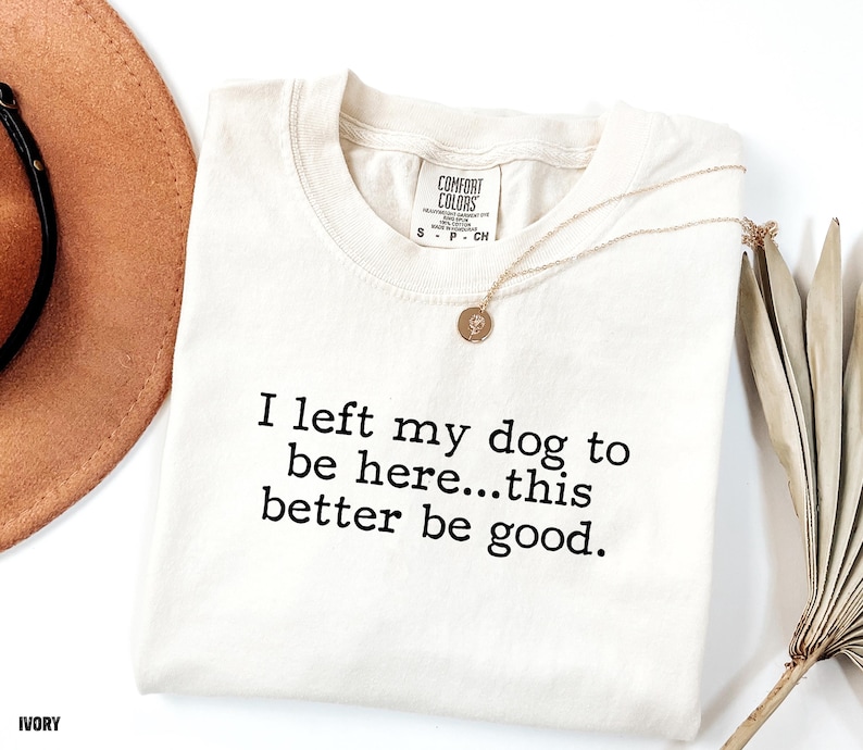 May include: Ivory-colored t-shirt with the text "I left my dog to be here...this better be good." A gold necklace with a pendant is on the shirt. A brown hat is in the background.