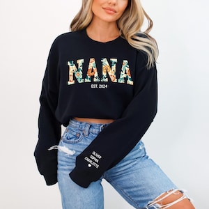 Op de afbeelding: Zwart gecropped sweatshirt met de tekst "NANA EST. 2024" in bloemenletters. Het sweatshirt heeft witte tekst op de mouw die "OLIVER, SOPHIA, CHARLOTTE" met een hartje zegt.