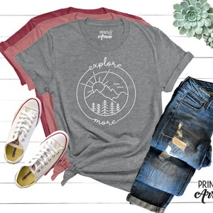 May include: Gray t-shirt with a white graphic of a mountain range, sun, and trees in a circle with the text "explore more". The shirt is laid out on a white surface with a pair of white sneakers, a pair of blue jeans, and two succulents.