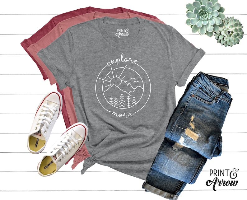 May include: Gray t-shirt with a white graphic of a mountain range, sun, and trees in a circle with the text "explore more". The shirt is laid out on a white surface with a pair of white sneakers, a pair of blue jeans, and two succulents.