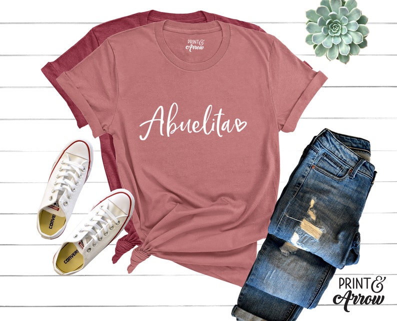May include: A pink t-shirt with the word "Abuelitas" written in white cursive with a small heart next to it. The shirt is folded and displayed on a white surface with a pair of white sneakers and blue jeans.