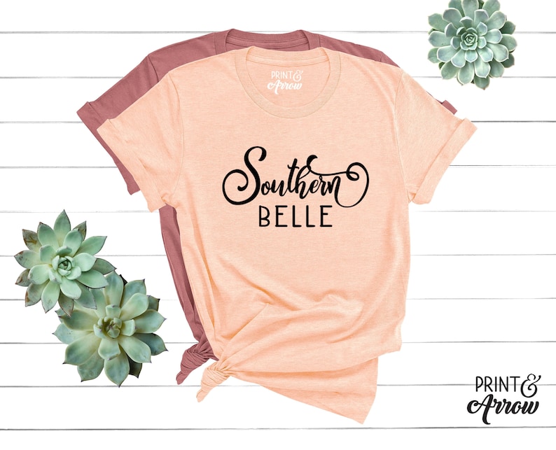 Puede incluir: Una camiseta de color melocot&oacute;n con el texto "Southern Belle" en fuente de letra cursiva negra. La camiseta est&aacute; doblada y se muestra sobre una superficie de madera blanca.