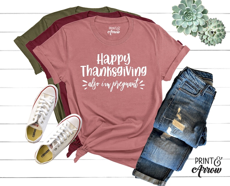 May include: A pink t-shirt with the text "Happy Thanksgiving also i'm pregnant" in white lettering. The shirt is laid out on a white surface with a pair of white trainers and a pair of blue jeans.