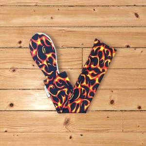 May include: A black and yellow flame pattern fabric arm warmer with a white trim. The arm warmer is folded in half and is lying on a wooden surface.