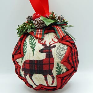May include: A round Christmas ornament with a red and black plaid pattern. The center features a deer design. The ornament is decorated with pinecones, greenery, and a red ribbon for hanging. Festive holiday decor.