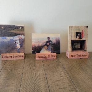 May include: Three wooden photo display stands with personalized text. The first stand has a photo of two people fishing and the text "Fishing Buddies". The second stand has a photo of a family with a dog and the text "November 21, 2015". The third stand has a photo of a person standing in a doorway and the text "Your Text Here".