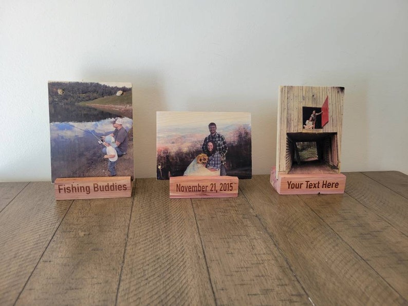 May include: Three wooden photo display stands with personalized text. The first stand has a photo of two people fishing and the text "Fishing Buddies". The second stand has a photo of a family with a dog and the text "November 21, 2015". The third stand has a photo of a person standing in a doorway and the text "Your Text Here".