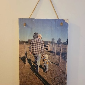 May include: A rustic wooden sign with a printed photo of a father and son walking in a field. The father is wearing a plaid shirt and a baseball cap, and the son is wearing a striped shirt. The photo is printed on a weathered wooden surface.