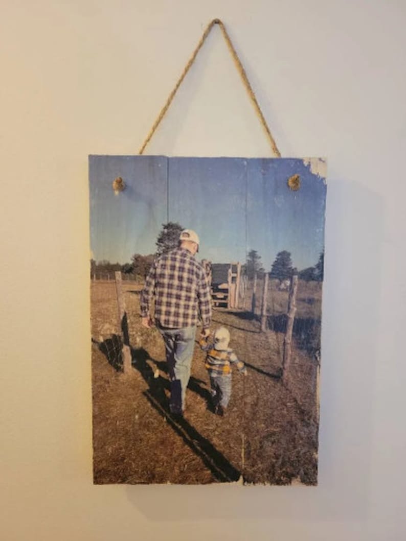May include: A rustic wooden sign with a printed photo of a father and son walking in a field. The father is wearing a plaid shirt and a baseball cap, and the son is wearing a striped shirt. The photo is printed on a weathered wooden surface.