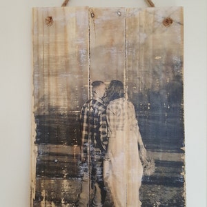 May include: A black and white photograph of a couple kissing, printed on a rustic wooden sign with a distressed finish. The sign is hanging from a twine rope.