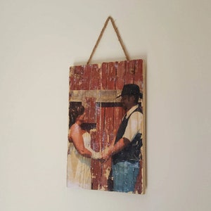 May include: A rustic wooden sign with a printed photo of a couple in front of a red barn. The sign has a distressed, weathered look and is hanging from a rope.
