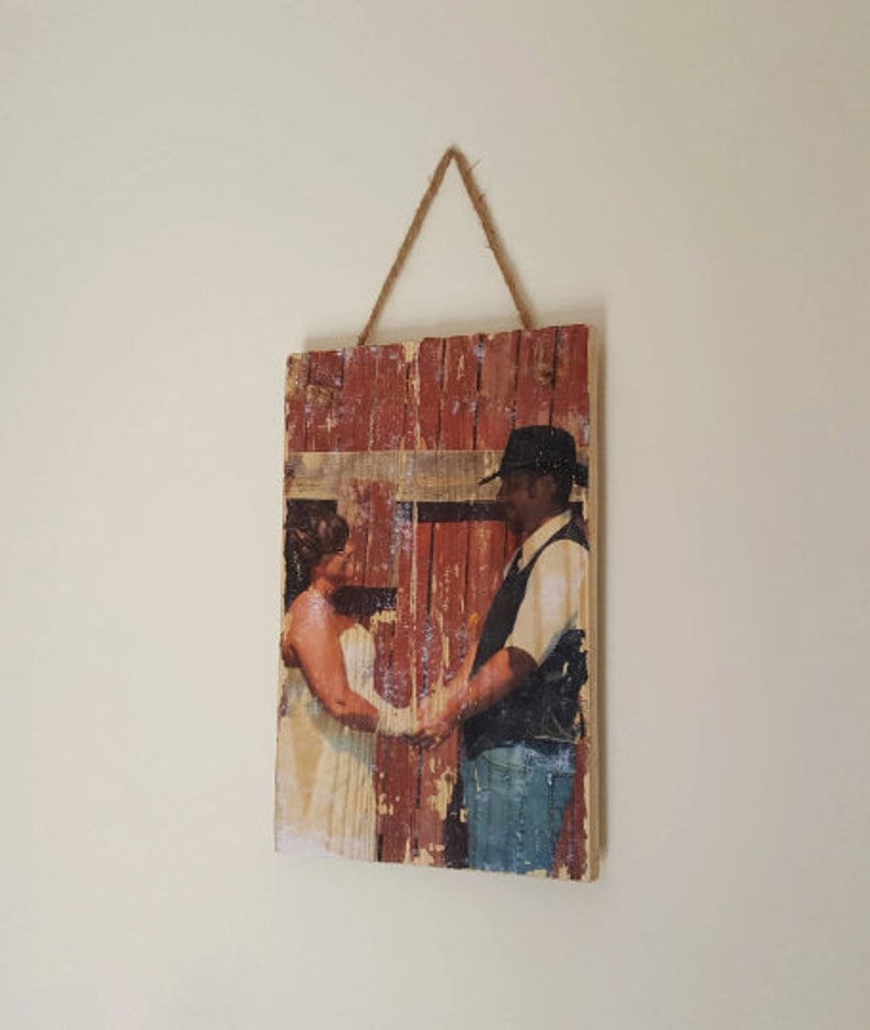 May include: A rustic wooden sign with a printed photo of a couple in front of a red barn. The sign has a distressed, weathered look and is hanging from a rope.