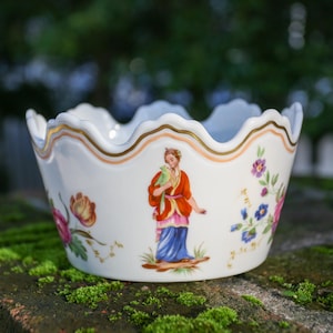Może przedstawiać: Biała porcelanowa miska z falistym brzegiem, ozdobiona kwiatowymi i figuralnymi ilustracjami. Miska ma złote obramowanie i centralny obraz osoby w tradycyjnym stroju. Miska jest ustawiona na rozmytym zielonym tle.