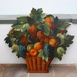 May include: A wooden wall hanging depicting a basket overflowing with painted fruit. The artwork features oranges, grapes, and apples, with green and brown leaves. The basket is a warm brown color, and the fruit is in various shades of orange, red, and purple.