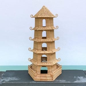 Puede incluir: Una pagoda decorativa de varios niveles hecha de mimbre tejido. La pagoda tiene un techo puntiagudo y ventanas abiertas en cada nivel. La estructura es de color marrón claro y se asienta sobre una superficie gris oscuro. La pagoda mide unos 61 cm de altura.