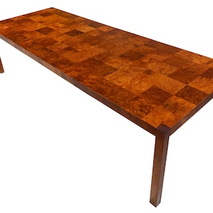 May include: A rectangular dining table featuring a tabletop with a patchwork pattern of square wooden tiles in varying shades of warm brown. The table is supported by four straight, slender legs that match the dark brown tone of the frame.