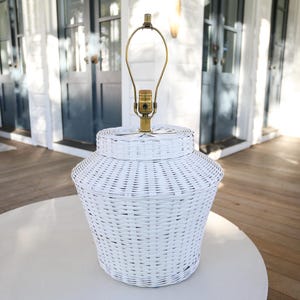 Op de afbeelding: Een witte rieten lamp met een messing hals en een witte kap. De lamp staat op een witte tafel op een houten terras. De lamp heeft een geweven mandontwerp.