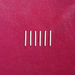 Peut inclure: Six bâtonnets blancs, fins et rectangulaires, disposés en rangée sur une surface rouge.