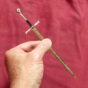 Könnte beinhalten: Ein kleines dekoratives Schwert mit goldenem Griff und Klinge, gehalten vor rotem Hintergrund. Das Schwert hat einen silbernen Parier und einen schwarzen Griff. Das Design erinnert an eine Fantasy-Waffe.