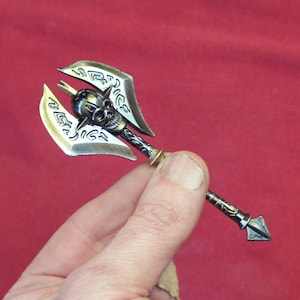 May include: A silver and gold axe keychain with a skull design and intricate details. The axe is held in a hand against a red background.