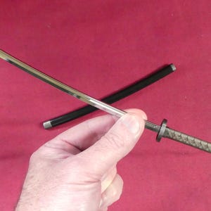 May include: A miniature katana sword with a silver blade and a black scabbard. The handle has a textured design. The sword is held against a red background.