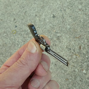 May include: A small, silver-coloured, toy revolver with a black handle. The toy gun is held in a hand.