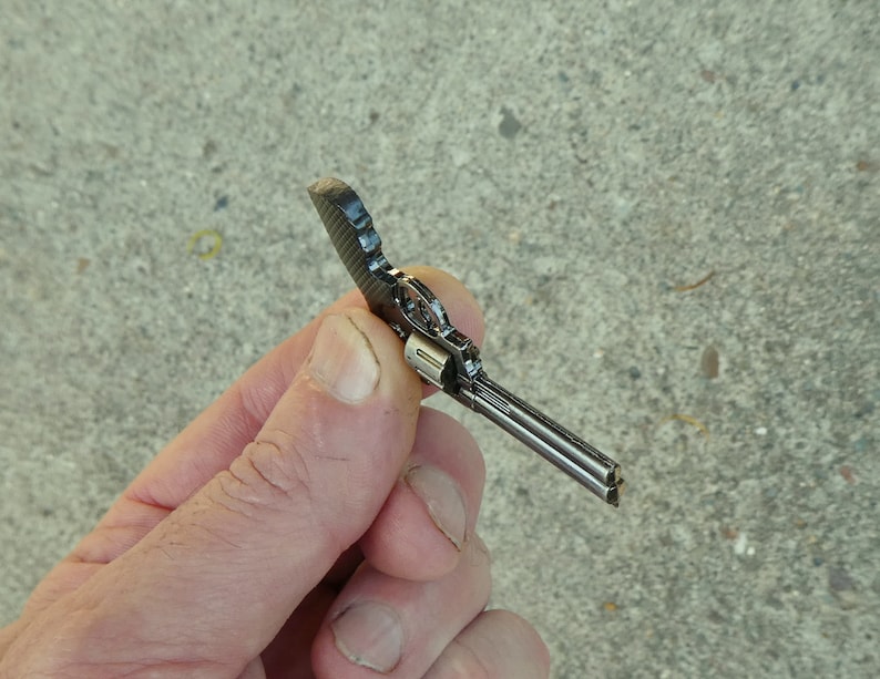 May include: A small, silver-coloured, toy revolver with a black handle. The toy gun is held in a hand.