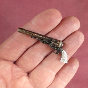 Könnte beinhalten: Miniatur-Revolver im antiken Stil mit aufwendigen Details. Die Pistole hat eine dunkle Metalloberfläche mit goldenen Akzenten und einen weißen Griff mit eingravierten Mustern. Kleiner Dekorationsartikel.